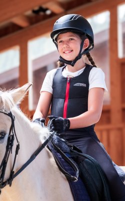Mädchen mit Helm und Schutzweste reitet ein weißes Pferd in einer Reithalle