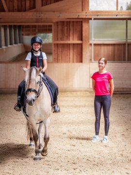 Kind reitet Pony in Reithalle, daneben steht Trainerin in rotem Shirt und schwarzen Leggings