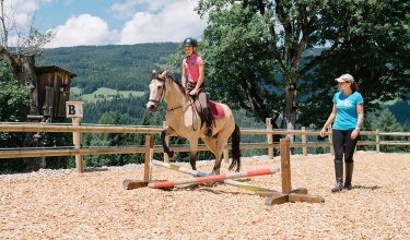 Mädchen reitet Pony über kleine Bodenstange, betreut von Reitlehrerin.