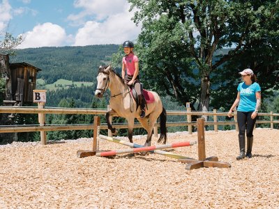 Mädchen reitet Pony über kleine Bodenstange, betreut von Reitlehrerin.