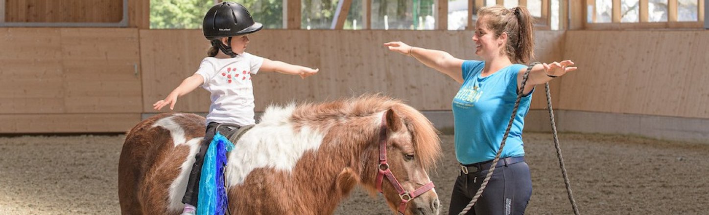 Kind übt Voltigierübung auf Pony unter Anleitung in der Reithalle.