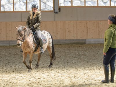 Kind reitet ein Pony in der Reithalle, begleitet von einer Trainerin.