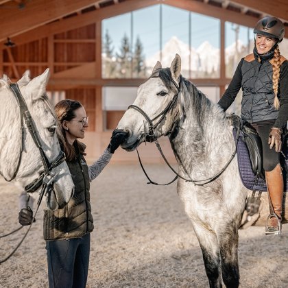 Frau streichelt Pferd, während eine Reiterin auf einem anderen Pferd in einer Reithalle sitzt