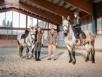 Zwei Menschen mit zwei Pferden in einer Reithalle, eine Frau sitzt auf einem Pferd, die andere steht daneben