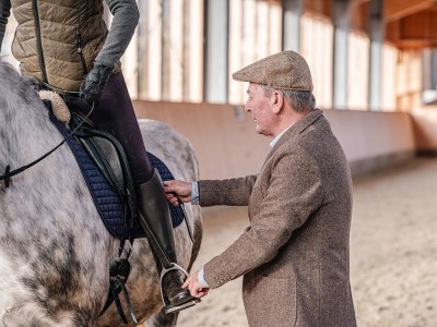 Älterer Mann hilft Reiterin beim Einsteigen in den Steigbügel eines Pferdes in einer Reithalle