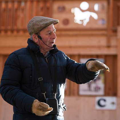 Älterer Mann mit Mütze und Mikrofon spricht und gestikuliert in einer Holzhalle