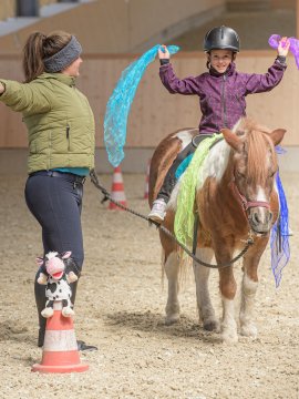 Kind balanciert fröhlich mit bunten Tüchern auf einem Pony beim Voltigieren.