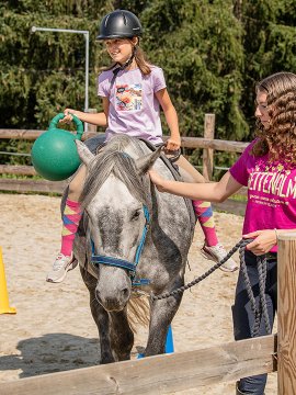 Mädchen zu Pferd hält grünen Ball im Parcours mit Begleitung.