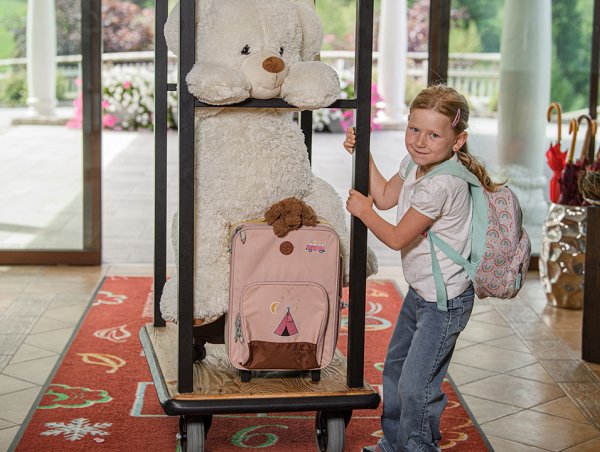 Mädchen mit Regenbogen-Rucksack hält Gepäckwagen mit großem Teddybär und rosa Koffer