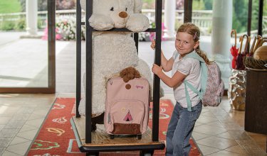 Girl with rainbow backpack holding luggage cart with large teddy bear and pink suitcase