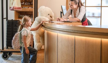 Child with backpack and large teddy bear at hotel reception, receptionist smiling