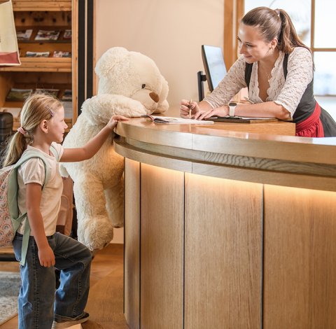 Kind mit Rucksack und großem Teddybär an der Hotelrezeption, Empfangsdame lächelt