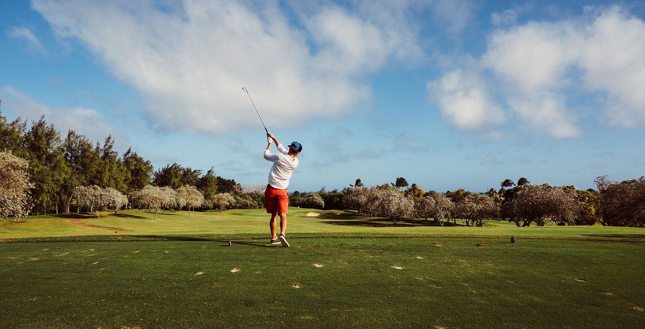 Golfer schlägt einen weiten Drive auf grünem Fairway unter blauem Himmel.