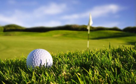 Golfball liegt im Gras nahe dem Green mit Fahne im Hintergrund.