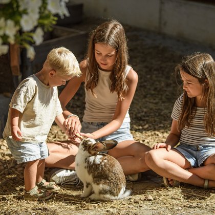 Drei Kinder sitzen und stehen auf Stroh, ein Kind füttert ein Kaninchen im Freien