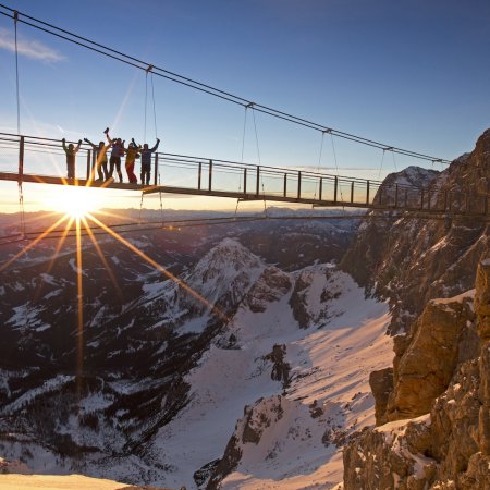 Gruppe auf Hängebrücke bei Sonnenuntergang mit verschneitem Bergpanorama.