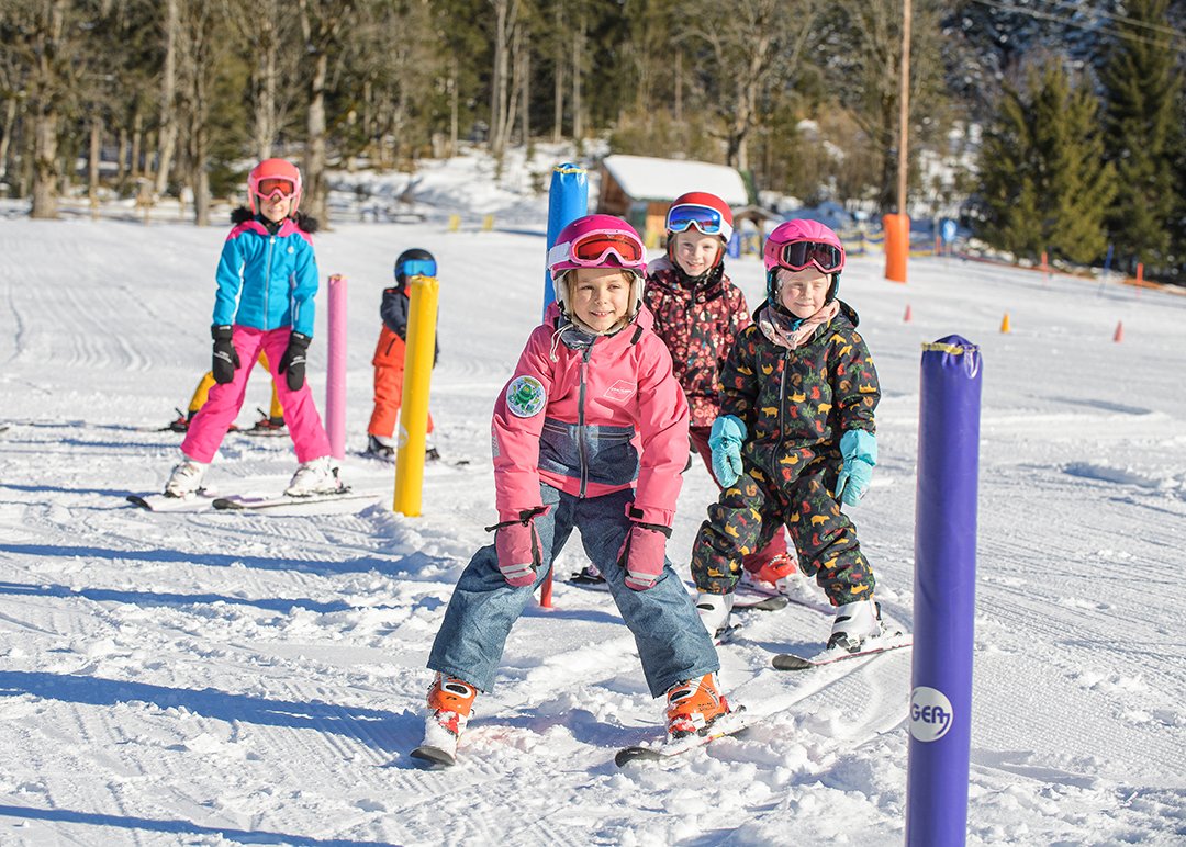 Kinder fahren bei Sonne durch bunte Stangen auf Übungspiste.