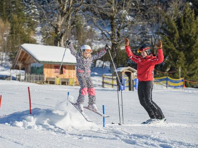 Mädchen springt über Schneehügel, Skilehrer jubelt daneben.