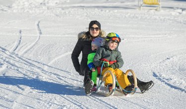 Mutter rodelt mit zwei Kindern eine verschneite Piste hinunter.