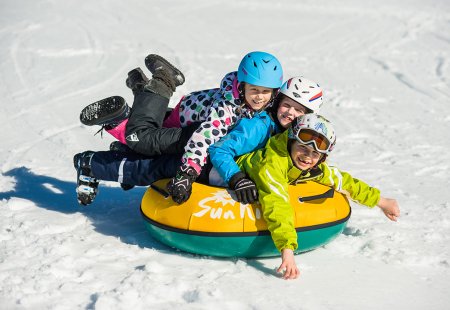 Drei fröhliche Kinder liegen auf einem Schneereifen übereinander.