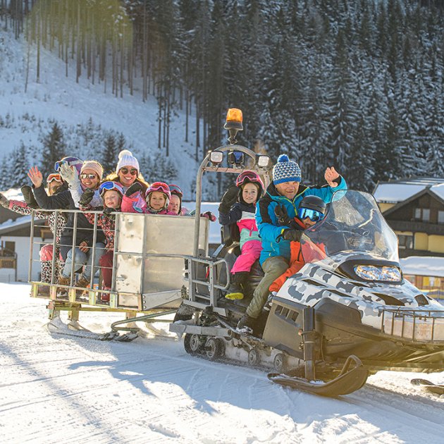 Schneemobilfahrt mit fröhlich winkenden Kindern und Erwachsenen im Winterpanorama.