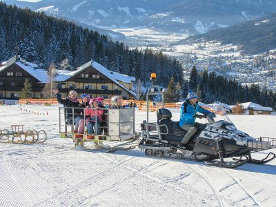 Schneemobil zieht Schlitten mit winkenden Kindern und Erwachsenen durch den Schnee.