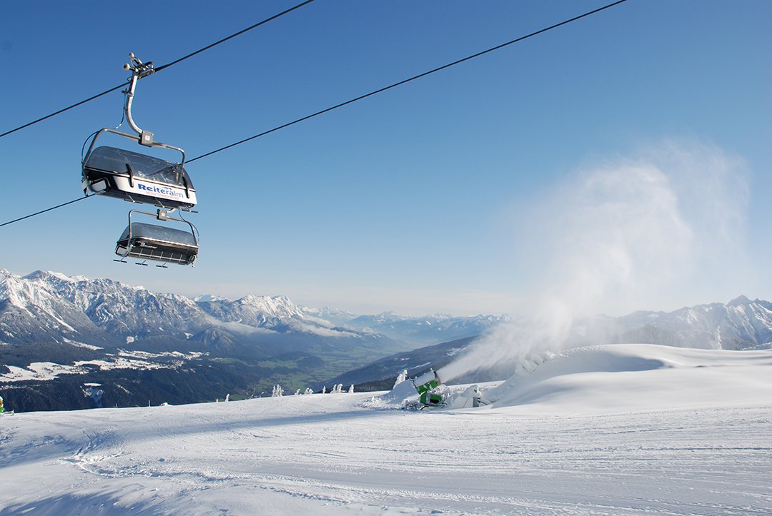 Sessellift über Pisten mit Schneekanone und grünem Tal im Hintergrund.